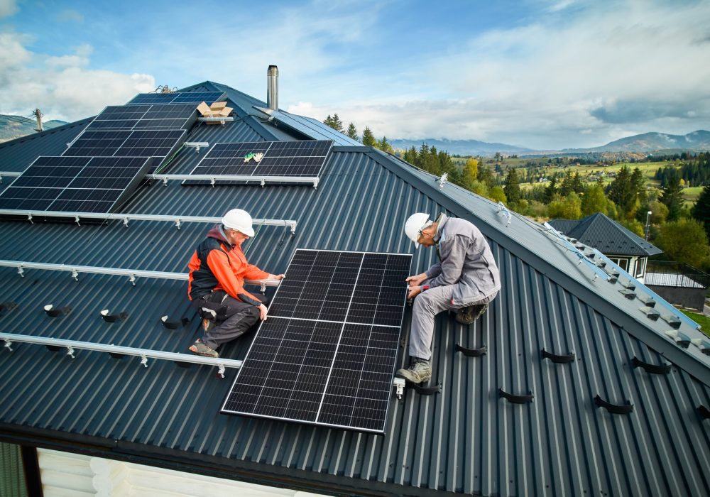 Roofers building photovoltaic solar module station on roof of house. Men electricians in helmets installing solar panel system outdoors. Concept of alternative and renewable energy. Aerial view.